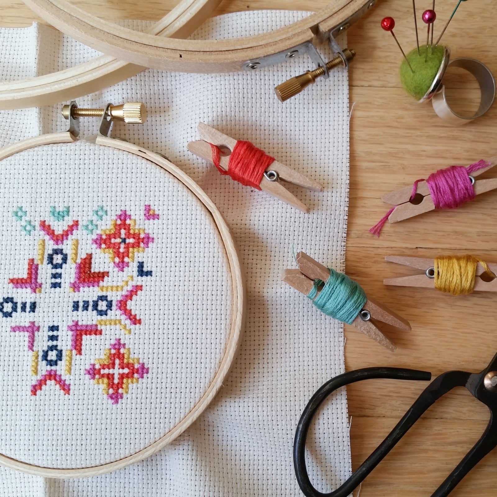 A geometric cross-stitch piece in a wooden hoop next to colorful thread wound around mini wooden clothespins.
