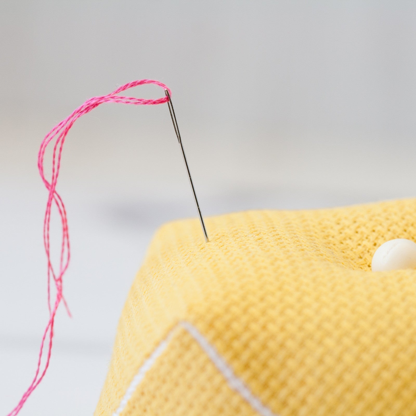 A close-up of a silver sewing needle threaded with pink floss, stuck into a yellow textured pincushion.