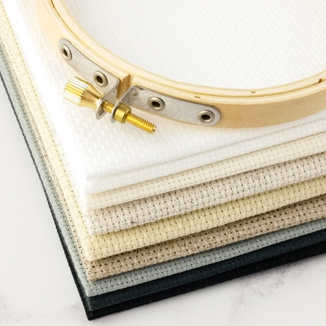 A wooden embroidery hoop resting on a stack of cross-stitch Aida fabrics ranging from white to black.
