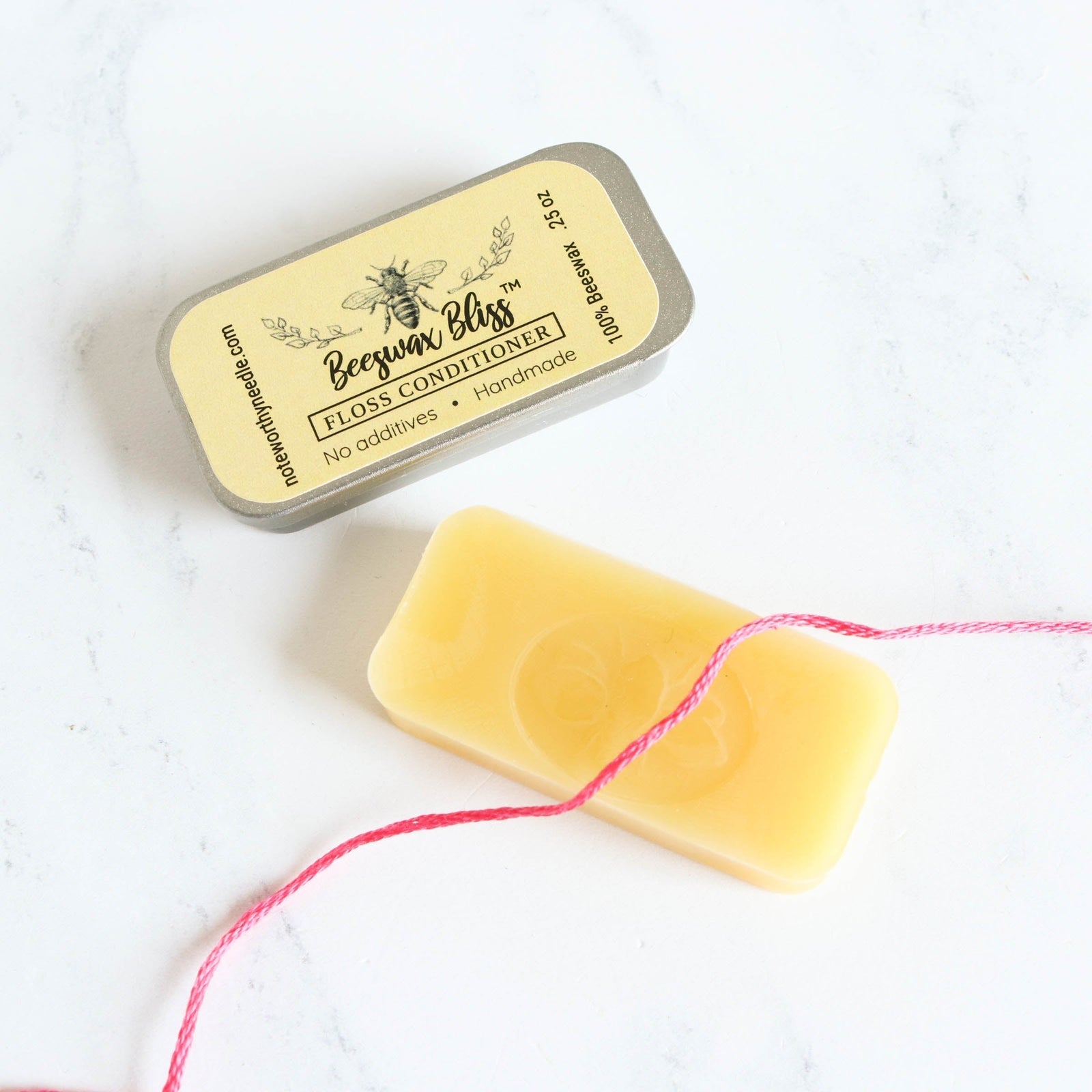 Beeswax Bliss floss conditioner bar with its tin and a strand of pink embroidery thread on white marble.