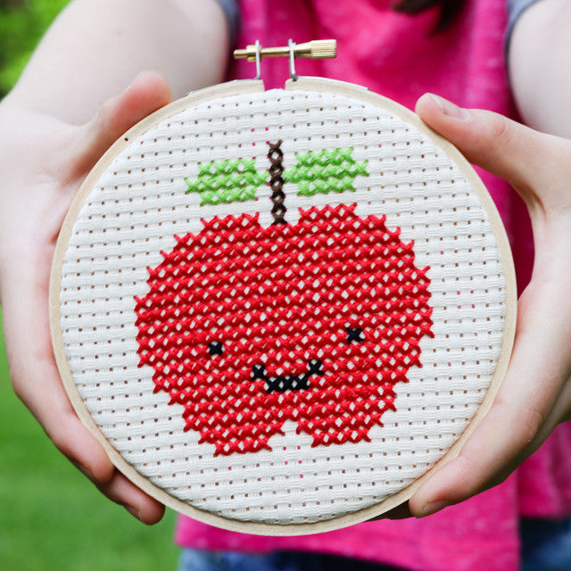 A person holding a wooden hoop displaying a finished cross-stitched smiling red apple.