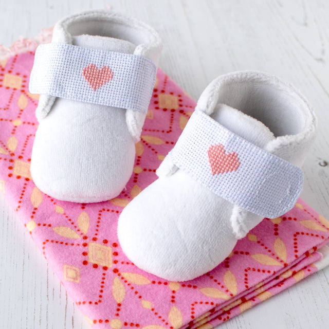 Pair of soft white baby booties with pink cross-stitch hearts on velcro straps, on a pink patterned blanket.