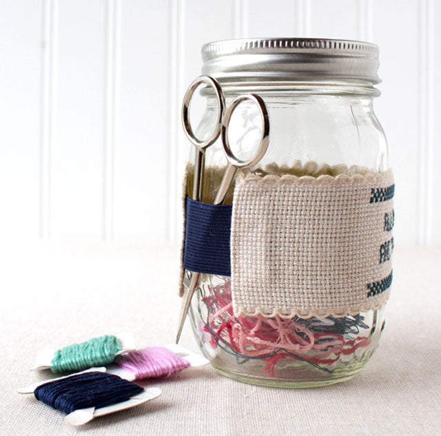 A glass mason jar wrapped in an embroidered cuff holding scissors, with thread bobbins nearby.