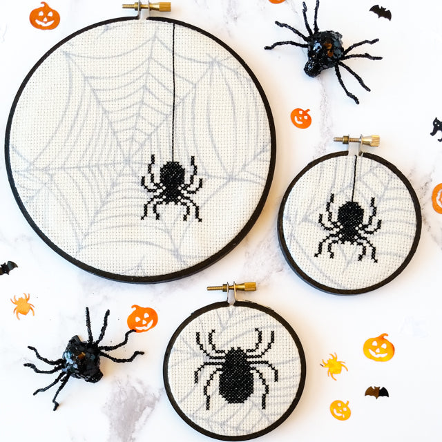 Three Halloween cross-stitch hoops featuring spider and web designs surrounded by toy spiders and pumpkin confetti.