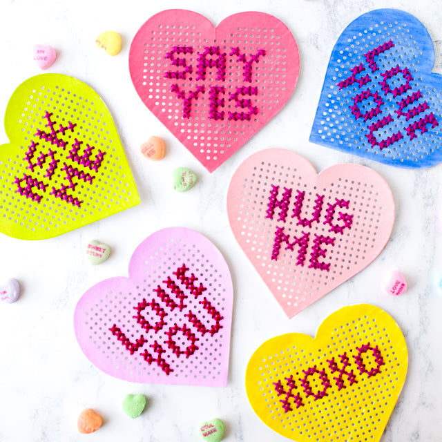 Cross-stitched perforated felt hearts with romantic phrases like "HUG ME" and "SAY YES" on marble.