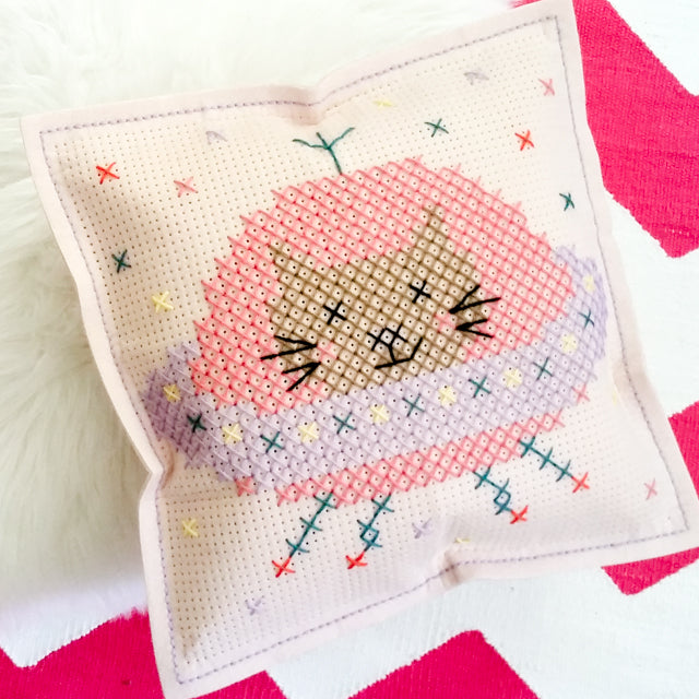 A finished cross-stitch pillow featuring a cat face inside a pink and purple flying saucer.