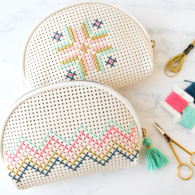 Two cream cross-stitched pouches with geometric patterns next to embroidery floss and gold scissors.