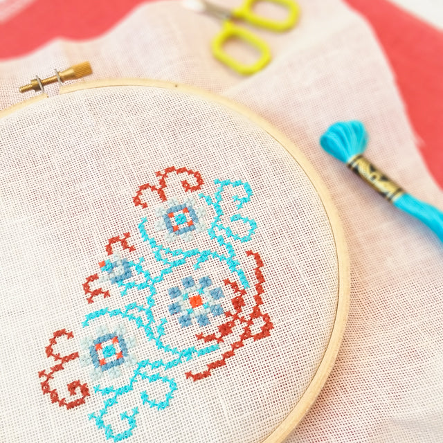 A wooden hoop framing an in-progress floral cross-stitch pattern alongside blue embroidery floss