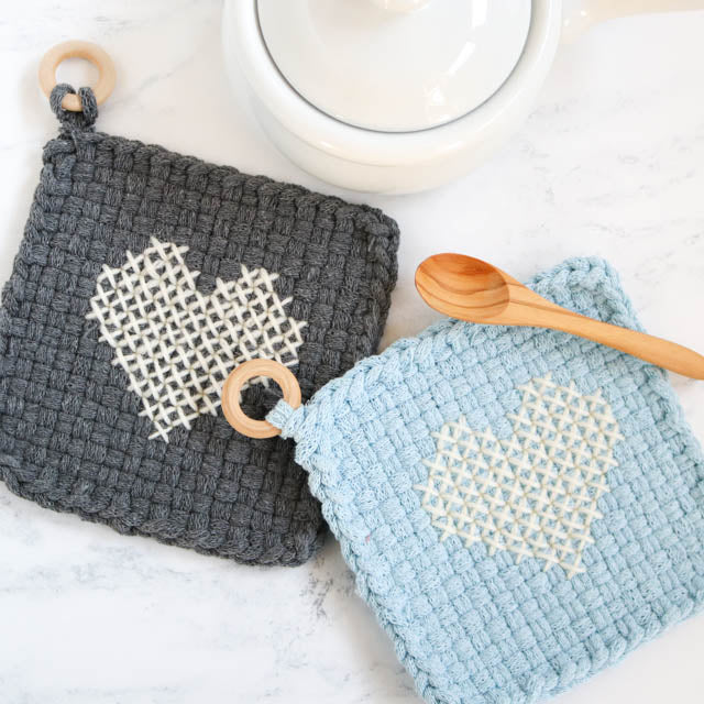Two crocheted heart-shaped potholders beside wooden cooking spoons on a light surface.