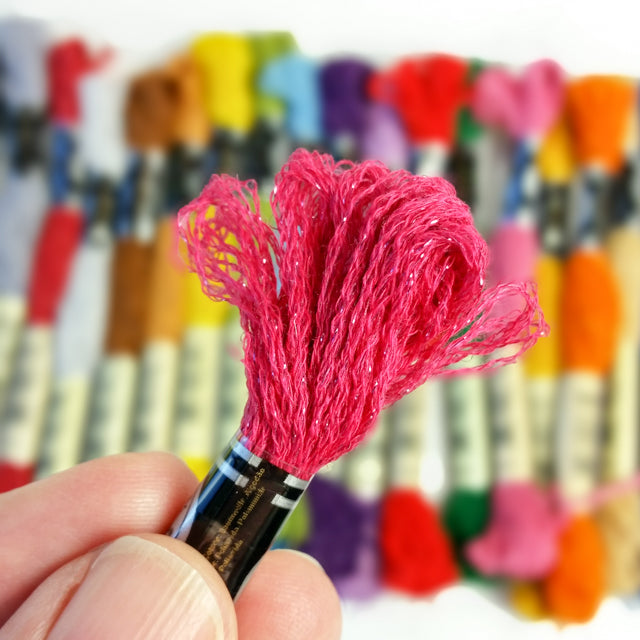A hand holding a sparkly pink embroidery floss skein against a blurred background of colorful threads.