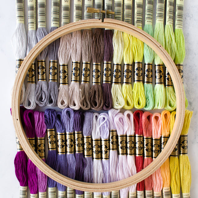 A wooden embroidery hoop placed over rows of colorful DMC embroidery floss skeins organized by shade.