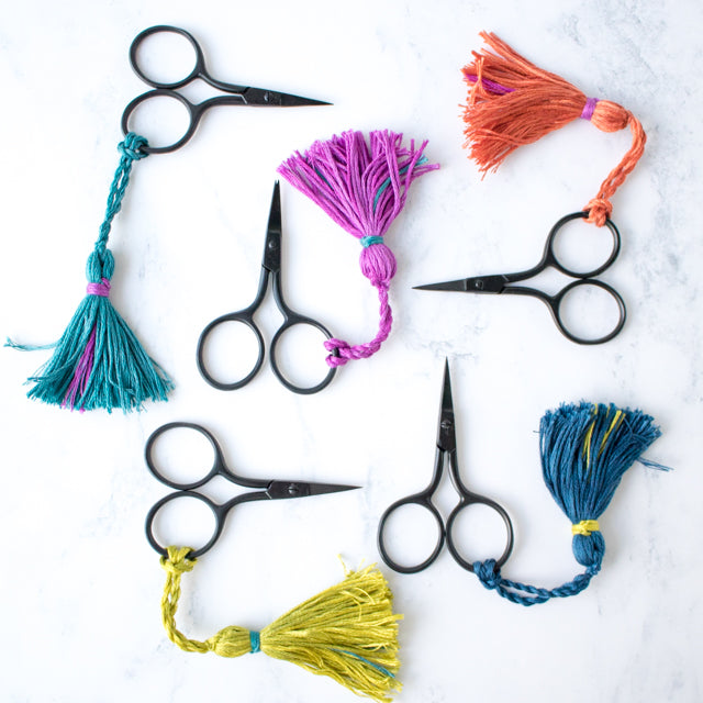 Five black embroidery scissors with colorful yarn tassels on a marble surface.