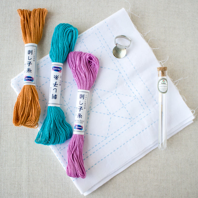 Three colorful sashiko thread skeins resting on white fabric with blue pattern lines, next to a thimble and needles.