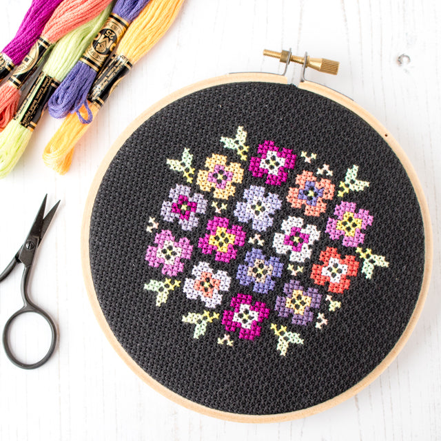 A stitched floral geometric design on black fabric in a wooden hoop, alongside bright embroidery floss skeins and scissors.