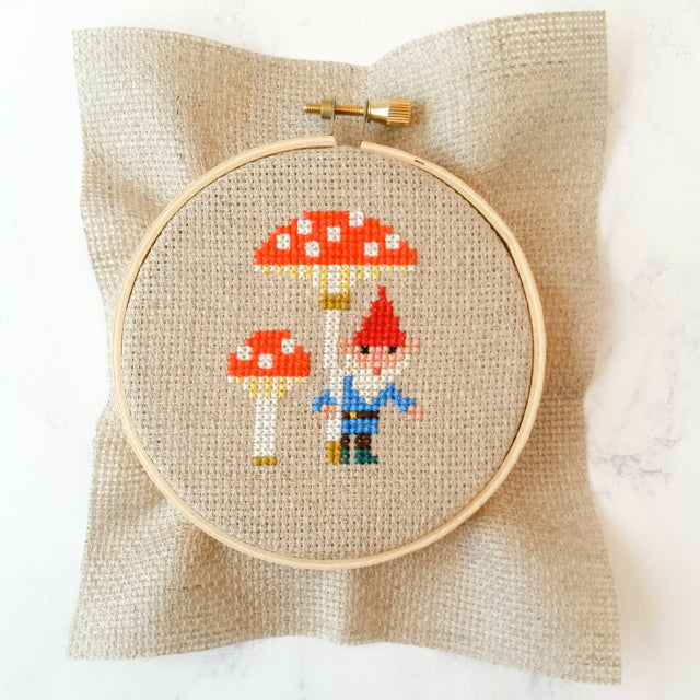Cross-stitch design of a garden gnome standing next to red mushrooms, secured inside a wooden embroidery hoop.