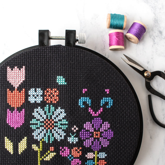 Floral cross-stitch pattern on black fabric in a hoop, next to three spools and scissors on marble.