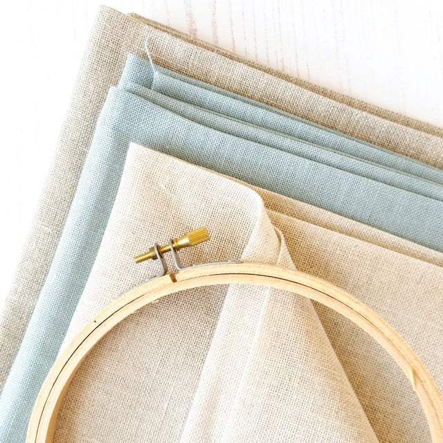 A wooden embroidery hoop resting on stacked linen fabrics in shades of cream, light blue, and tan.