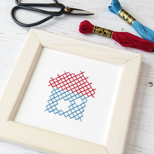A red and blue cross-stitched house pattern framed in light wood with embroidery floss and scissors nearby.