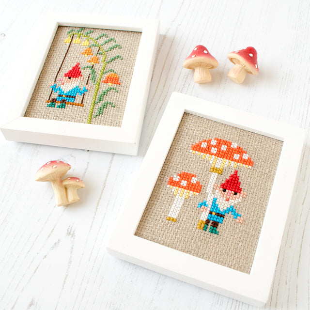 Two framed cross-stitch artworks featuring garden gnomes and mushrooms next to small mushroom figurines.