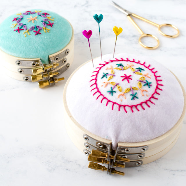 Two embroidered pincushions made from stacked embroidery hoops alongside heart-headed pins and scissors.