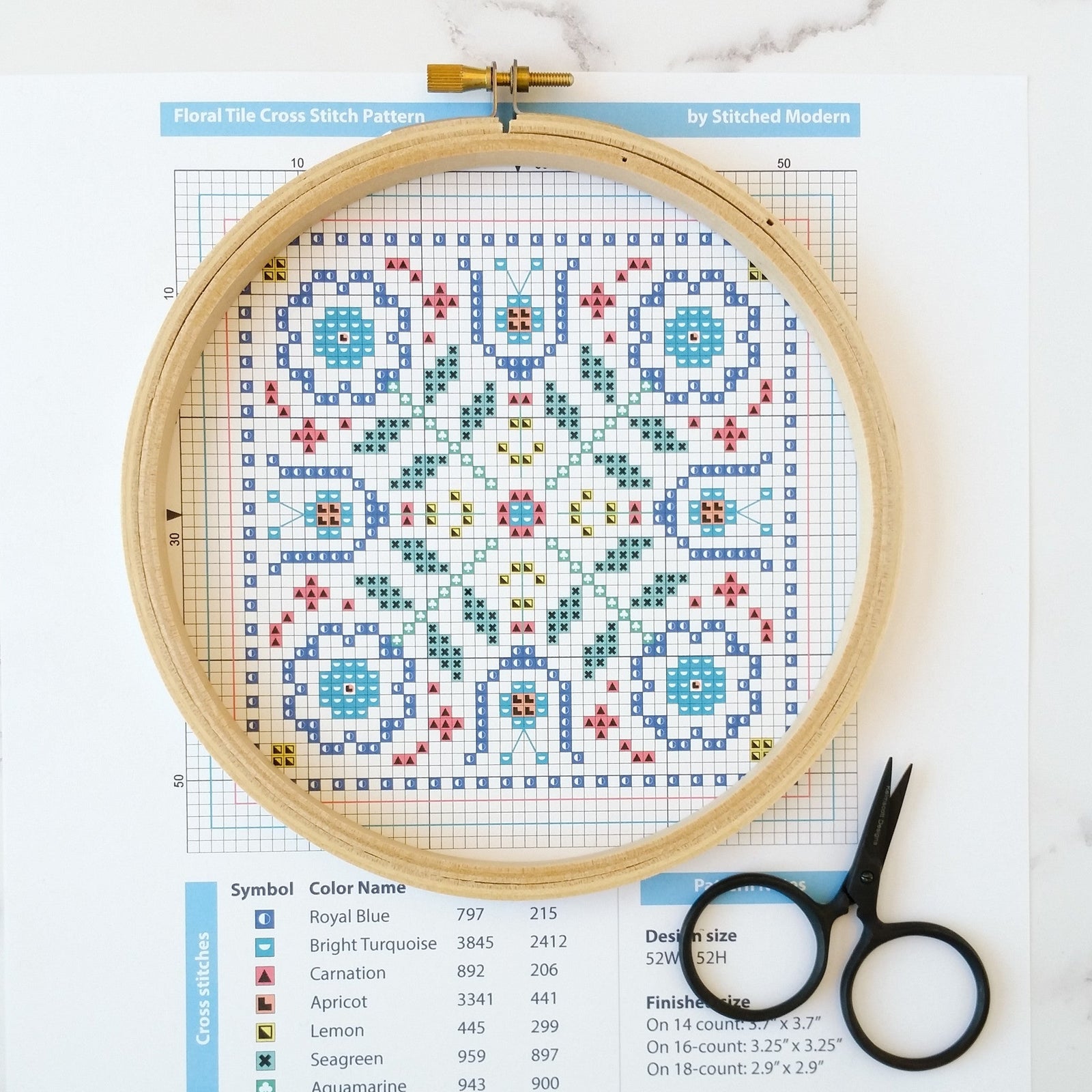 A wooden embroidery hoop framing a colorful floral cross-stitch pattern chart next to black scissors.