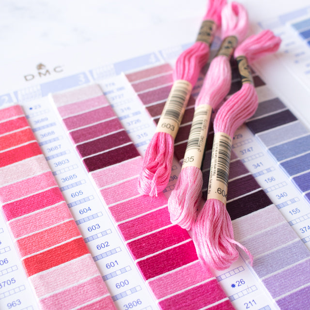 Three skeins of pink embroidery floss resting on top of a DMC color chart with various shades.