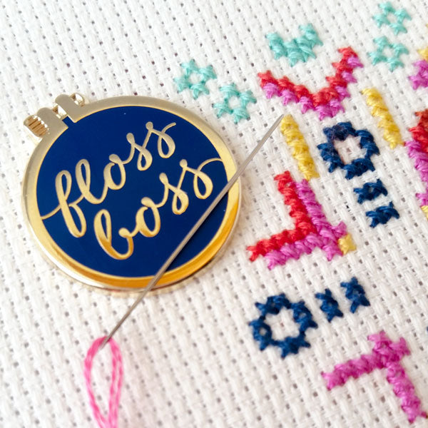 A blue enamel "floss boss" needle minder holding a needle on colorful cross-stitch fabric.