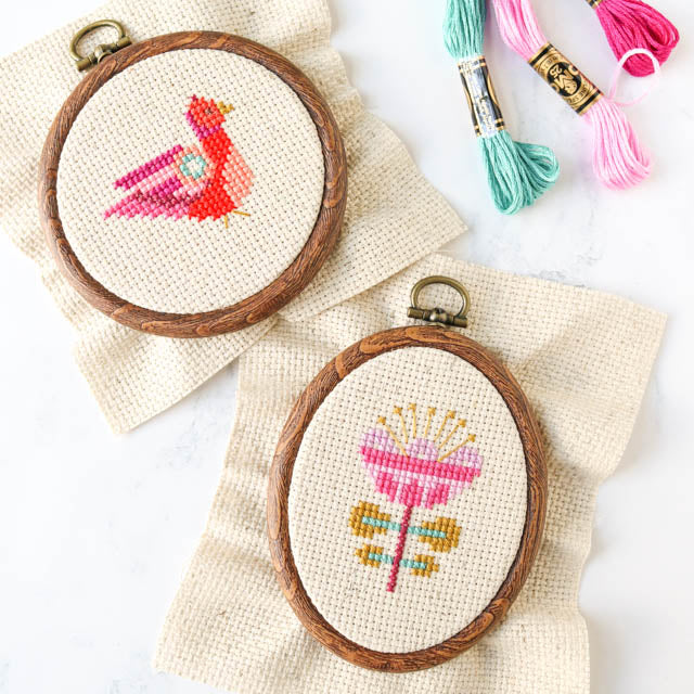 Two cross-stitched hoop designs of a colorful bird and pink flower with embroidery threads nearby.