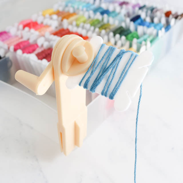 Plastic embroidery floss bobbin winder tool with blue thread clamped to an organizer box filled with bobbins.