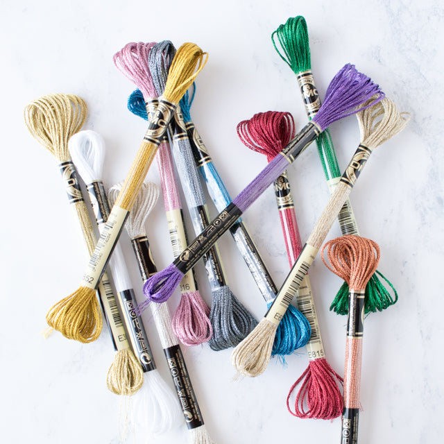 A colorful assortment of metallic and satin embroidery floss skeins scattered on a white marble background.