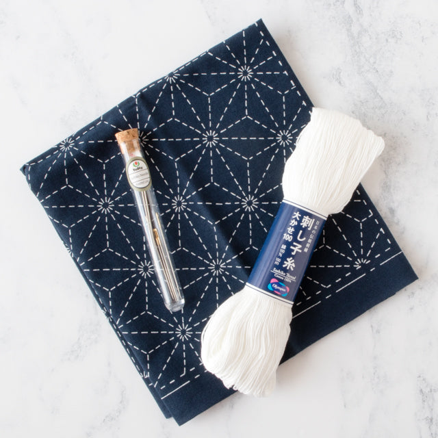 White sashiko thread and a tube of needles resting on a folded navy blue geometric starburst pattern fabric on marble.