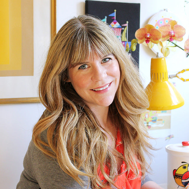 Person with long wavy blonde hair wearing a gray sweater and orange top, seated indoors near a lamp and orchids.
