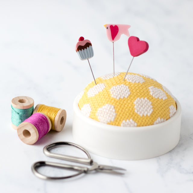 Yellow patterned pincushion with novelty pins shaped like a cupcake, bird, and heart, next to spools and scissors.