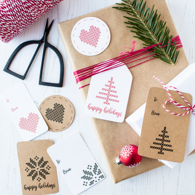 A wrapped gift decorated with a pine branch alongside holiday tags featuring cross-stitch holiday prints and scissors.