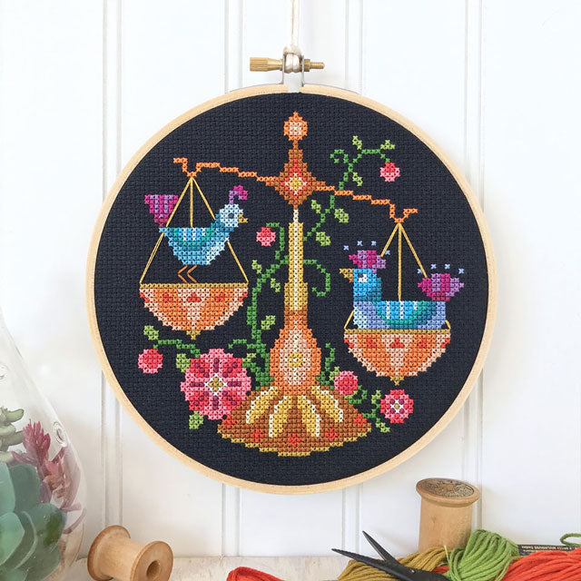 Cross-stitch art in a hoop depicting Libra scales with birds on black fabric, with spools and thread below.