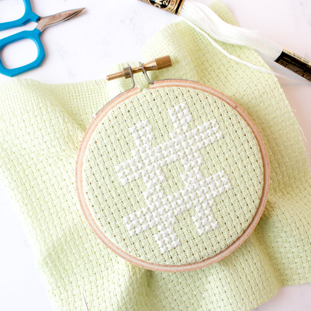 White cross-stitch hashtag design in a wooden hoop on light green fabric, with blue scissors and white floss skein.
