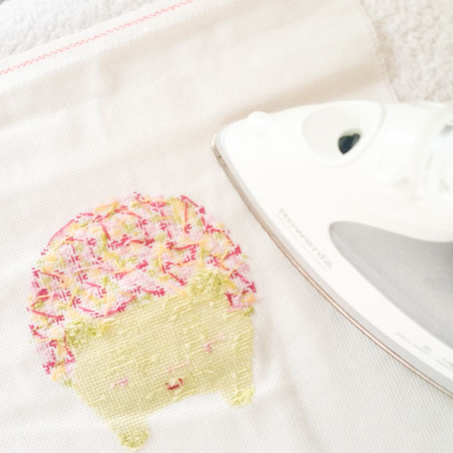 An iron pressing the reverse side of a cross-stitched character fabric to flatten the stitches.