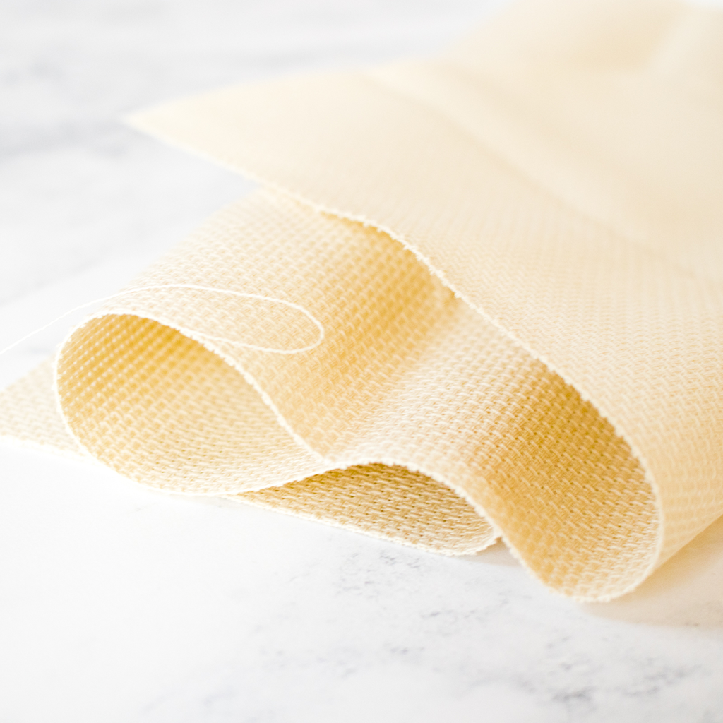 Soft cream-colored Aida fabric loosely folded on a white marble surface for a cross-stitch project.