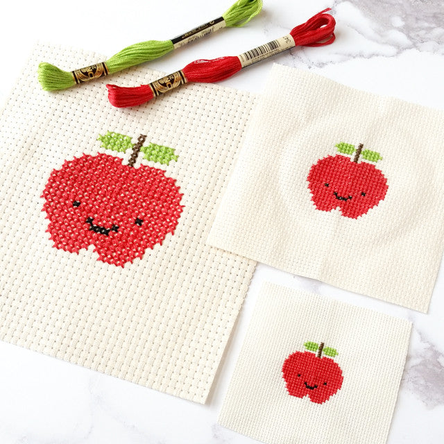 Three cross-stitched smiling red apples on different fabric counts with red and green floss skeins.