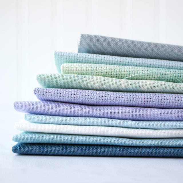 A folded stack of cross-stitch fabrics in cool pastel shades of blue, green, and purple.