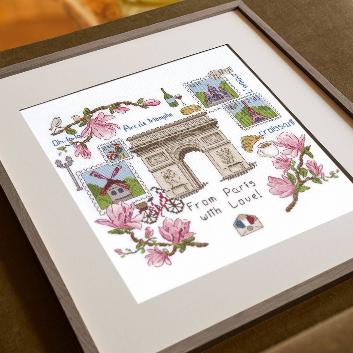 Framed cross-stitched artwork of the Arc de Triomphe with floral elements on a table.