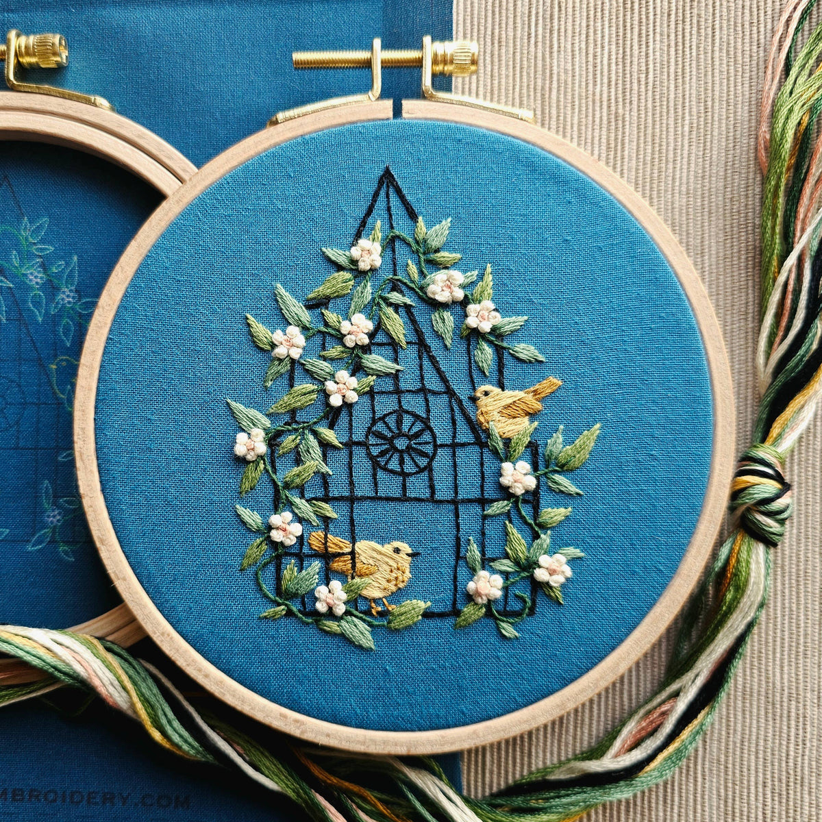 Hand embroidery project featuring a greenhouse trellis with climbing white jasmine flowers and birds on blue linen.