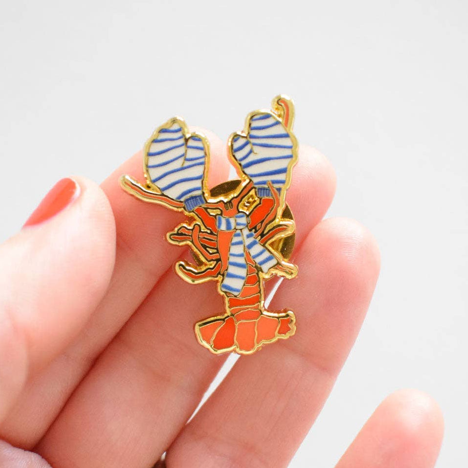 Enamel pin of an orange lobster wearing blue and white striped mittens and a matching scarf, held in a hand.
